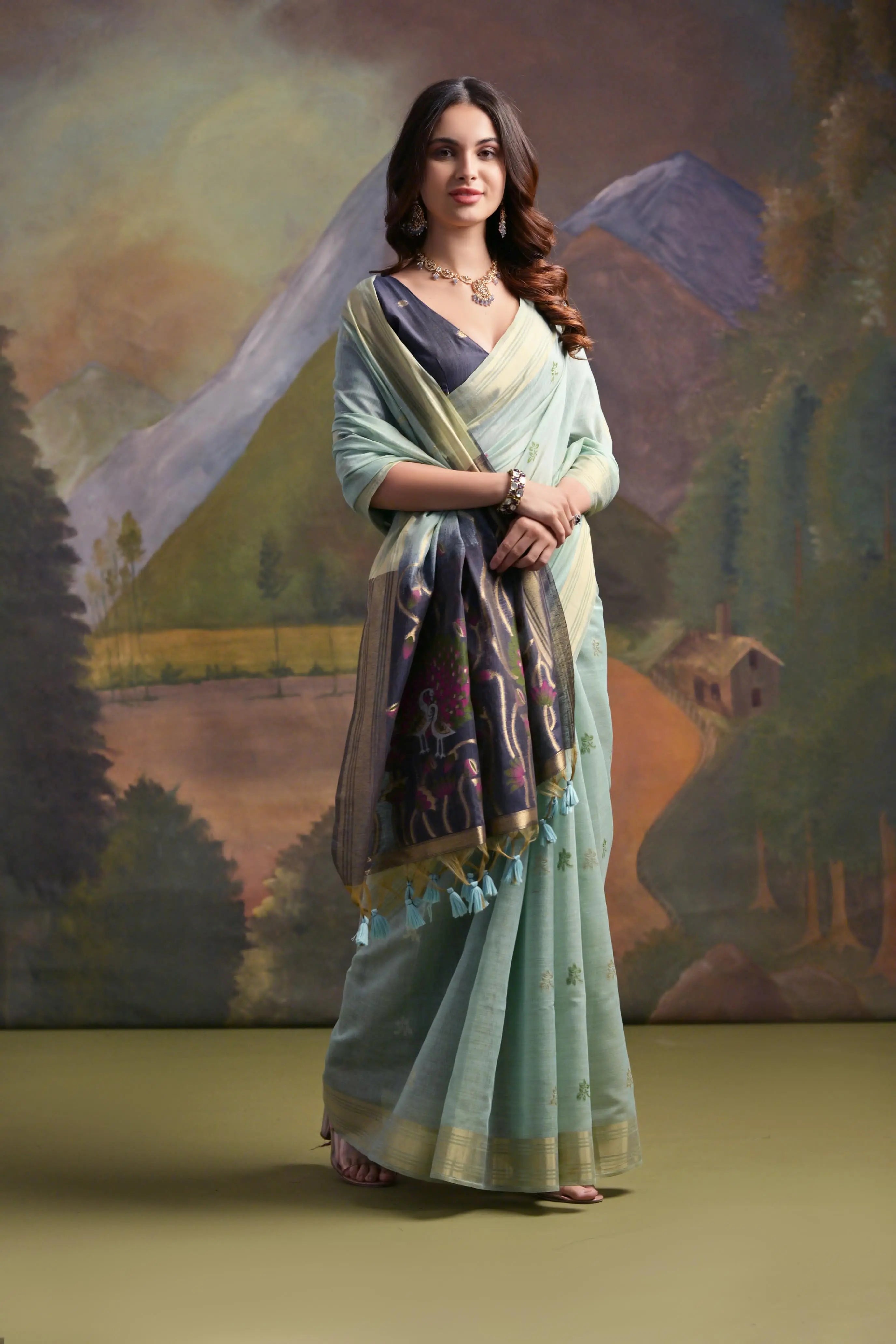 A woman models a soft Sea Green Muga Silk Saree with subtle Resham weaving. The pallu dramatically transitions to a deep indigo/black color, featuring intricate purple and green floral Resham threadwork. She wears a dark blouse and an oxidized silver choker necklace.